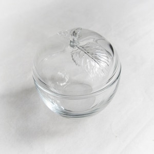 Può includere: Una ciotola di vetro trasparente con coperchio abbinato. Il coperchio presenta un motivo decorativo a foglia. La ciotola è rotonda e poggia su una superficie bianca. Ideale per servire o conservare.