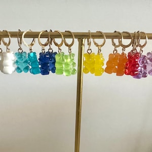 Può includere: Un set di 14 orecchini con ciondolo a forma di orsacchiotto gommoso in vari colori, tra cui nero, bianco, blu, verde, giallo, arancione, rosso, viola e rosa. Gli orecchini sono esposti su un supporto dorato.