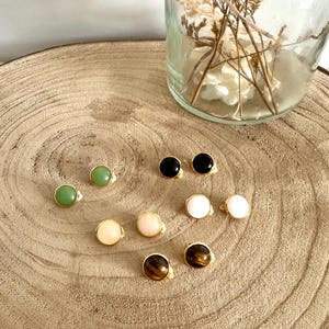 Peut inclure: Une collection de cinq paires de boucles d'oreilles à clous rondes avec des pierres cabochons lisses et polies en vert, noir, blanc, rose pâle et œil-de-tigre brun, chacune sertie dans une monture en métal doré sur une surface en bois.