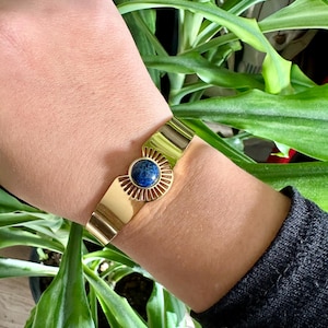 May include: A gold cuff bracelet with a blue gemstone in the center. The bracelet has a sunburst design around the gemstone.