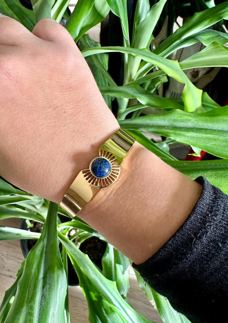 May include: A gold cuff bracelet with a blue gemstone in the center. The bracelet has a sunburst design around the gemstone.