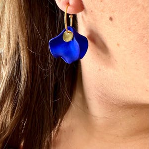 Op de afbeelding: Een paar gouden creolen met een blauwe bloemenhanger. De bloemenhanger is gemaakt van een zacht, buigzaam materiaal en heeft een licht gekrulde textuur.