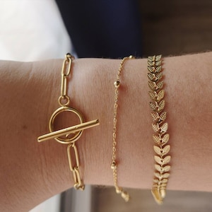 May include: Three gold bracelets on a wrist. One bracelet has a toggle clasp, one has a chain with small beads, and one has a chain of leaves.