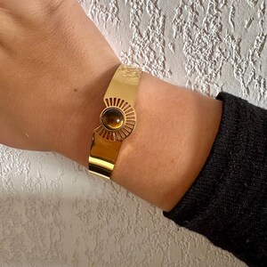 May include: A gold bangle bracelet with a sunburst design and a brown tiger's eye stone in the center.