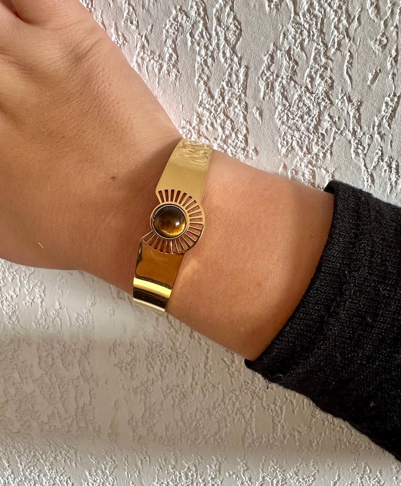 May include: A gold bangle bracelet with a sunburst design and a brown tiger's eye stone in the center.