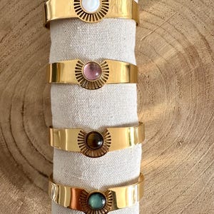 May include: Four gold bracelets with different colored stones in the center. The stones are white, pink, brown, and green.