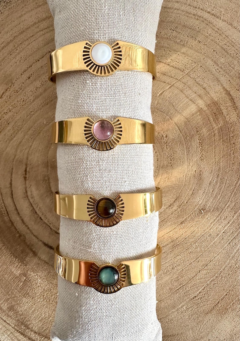 May include: Four gold bracelets with different colored stones in the center. The stones are white, pink, brown, and green.