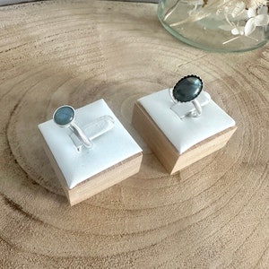 May include: Two silver rings with oval-shaped gemstones on wooden display stands. The ring on the left has a light blue gemstone, while the ring on the right has a dark grey gemstone.