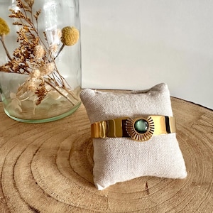 May include: A gold bangle bracelet with a green gemstone and a sunburst design. The bracelet is displayed on a small white cushion.