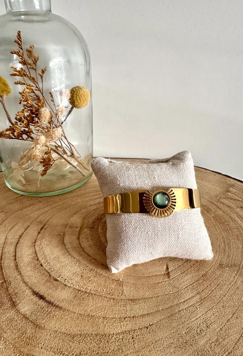 May include: A gold bangle bracelet with a green gemstone and a sunburst design. The bracelet is displayed on a small white cushion.