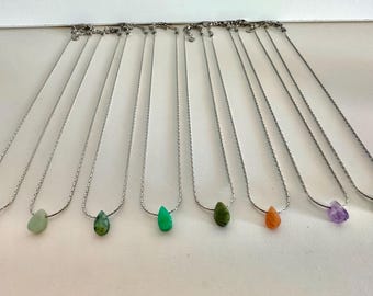 May include: A collection of silver necklaces, each featuring a teardrop-shaped gemstone pendant in various colors. The gemstones include blue, green, orange, and purple. The necklaces are displayed on a white surface.