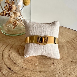 May include: A gold bracelet with a tiger's eye stone in the center. The bracelet is on a small white pillow.