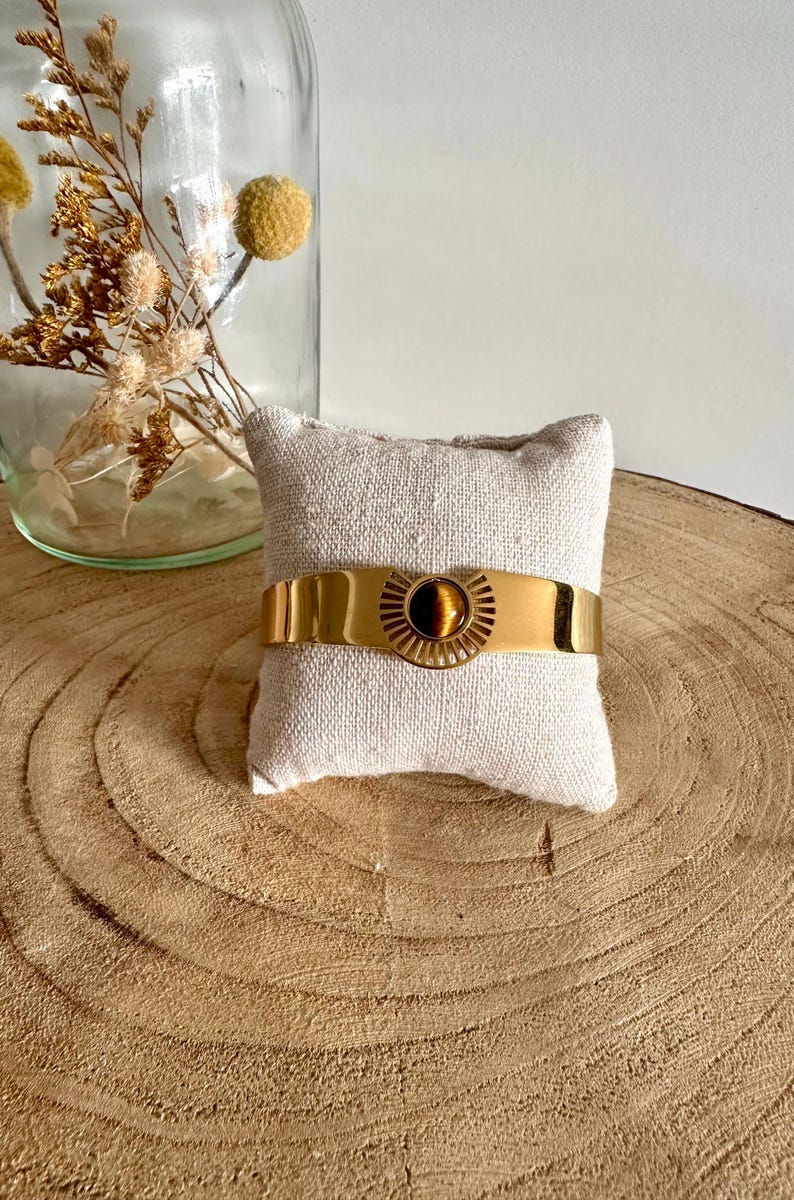 May include: A gold bracelet with a tiger's eye stone in the center. The bracelet is on a small white pillow.