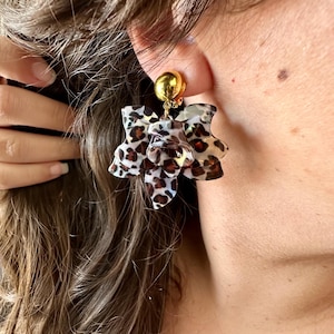 Peut inclure: Boucles d'oreilles à clous dorées avec un motif floral. Les pétales de la fleur présentent un imprimé léopard dans les tons marron, noir et crème. Les boucles d'oreilles sont portées à l'oreille d'une personne.
