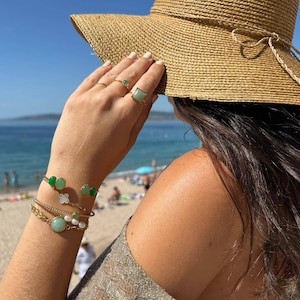 Puede incluir: Una mujer lleva un sombrero de paja y varias pulseras de oro con piedras verdes y blancas. Las pulseras están apiladas en su muñeca.