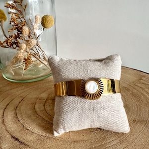 May include: A gold cuff bracelet with a white mother-of-pearl sunburst design. The bracelet is displayed on a small beige cushion.