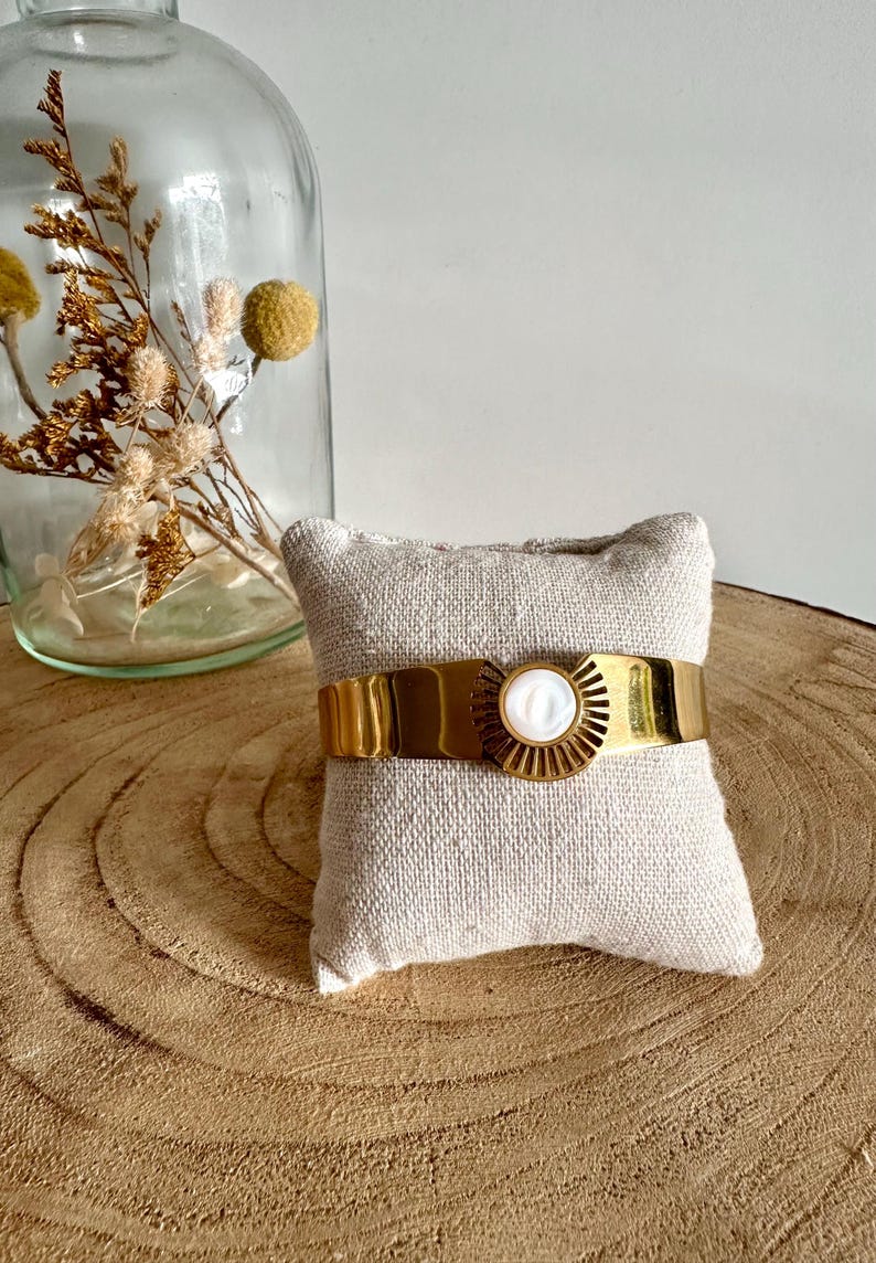 May include: A gold cuff bracelet with a white mother-of-pearl sunburst design. The bracelet is displayed on a small beige cushion.