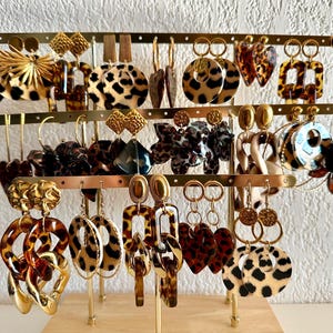 Può includere: Esposizione di vari orecchini in una gamma di stili e colori. Gli orecchini includono design in oro, tartaruga e stampa leopardata. L'esposizione è su un supporto color oro con una base in legno. Gli orecchini sono un mix di cerchi, pendenti e borchie.