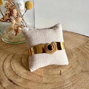 May include: A gold bangle bracelet with a tiger's eye stone set in a sunburst design. The bracelet is displayed on a small white pillow.
