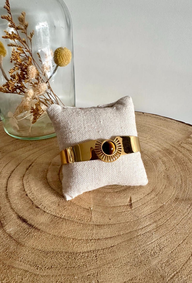 May include: A gold bangle bracelet with a tiger's eye stone set in a sunburst design. The bracelet is displayed on a small white pillow.