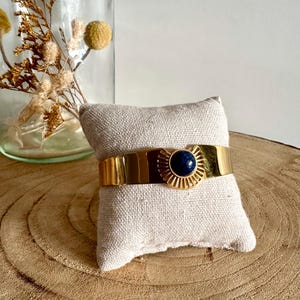 May include: A gold cuff bracelet with a blue gemstone in the center. The bracelet has a sunburst design around the gemstone.