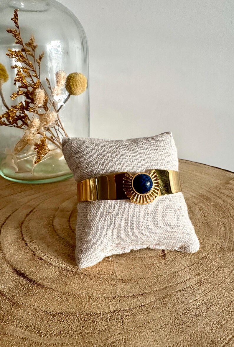 May include: A gold cuff bracelet with a blue gemstone in the center. The bracelet has a sunburst design around the gemstone.