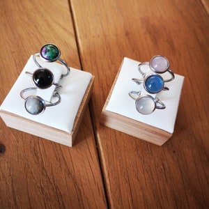 Peut inclure: Quatre bagues en argent avec des pierres précieuses de différentes couleurs. Les bagues sont empilées sur deux présentoirs blancs. Les présentoirs sont en bois et ont une finition brun clair.