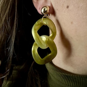 Peut inclure: Boucles d'oreilles vertes olive en forme de maillons avec un clou doré. Les boucles d'oreilles sont faites d'un matériau translucide et ont un design en forme de chaîne. Les boucles d'oreilles sont portées à l'oreille.