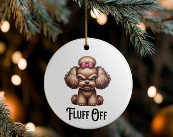 May include: A white, round Christmas ornament with a cartoon illustration of a brown poodle with an angry expression and a pink bow. The text "FLUFF OFF" is below the dog. The ornament hangs from a gold string.