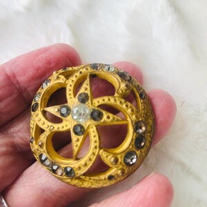 May include: A round, antique-style button with a gold finish. The button features a star design in the center, surrounded by a decorative pattern and embellished with small, sparkling rhinestones. The button is approximately 1.5 inches in diameter.
