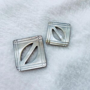 May include: Two square, silver-toned buttons with an oval cutout in the center. The buttons have a textured, grid-like design around the edges and a pearlescent sheen. The buttons are displayed on a soft, white surface.