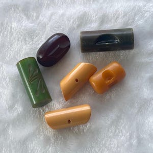 May include: A collection of vintage, colorful Bakelite buttons in various shapes. The buttons include a dark red oval, a green cylinder with etched design, and several tan and brown cylindrical buttons. The buttons are arranged on a white, textured surface.