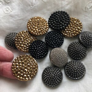 May include: Assortment of round, decorative buttons in gold, silver, and black. The buttons have a textured, beaded surface.