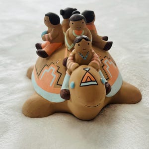 May include: A ceramic turtle figurine with five small figures riding on its back. The turtle is tan with orange and blue accents. The figures are wearing orange and tan clothing.