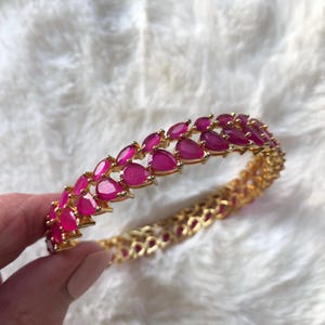 May include: Gold-toned bracelet with a repeating pattern of teardrop-shaped, magenta-colored gemstones. The bracelet is held against a white, textured background, showcasing the jewelry's intricate design and vibrant color.
