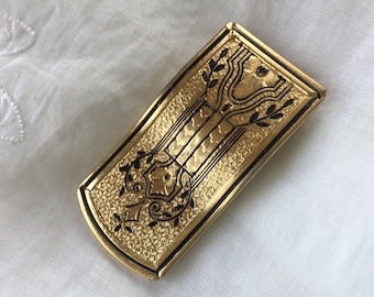 May include: A gold-colored metal brooch with a black design of stylized leaves and a central motif. The brooch has a textured surface.