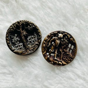 May include: Two antique brass buttons. One features a windmill and building scene, the other depicts a figure in a forest setting. Both buttons have a detailed, raised relief design and a dark, aged patina.