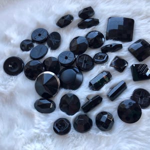 May include: A collection of black glass buttons in various shapes and sizes. The buttons are arranged on a white fuzzy surface.