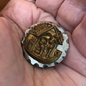 May include: A gold-toned metal button with a raised image of a house with two figures inside. The button has a silver-toned border with a scalloped edge.