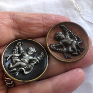 May include: Two antique metal buttons featuring cherub figures holding wreaths. The buttons are circular and have a raised design. One button has a dark brown background and the other has a light brown background.