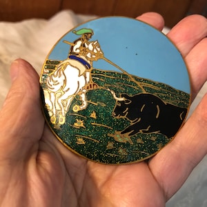 May include: A round, gold-toned metal pin with a blue, green, white, and black enamel design. The design depicts a man on a white horse with a spear, chasing a black bull.