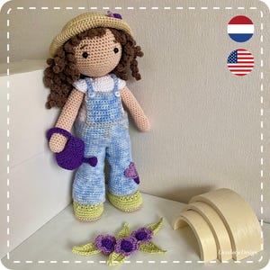 Op de afbeelding: Een gehaakte pop met een strohoed en een blauwe overall houdt een paarse gieter vast. De pop staat op een wit oppervlak met een houten boog op de achtergrond.