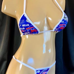 Puede incluir: Conjunto de bikini azul y blanco con el logotipo de los Buffalo Bills. La parte superior triangular y la parte inferior de tanga tienen correas y ribetes blancos. La tela muestra el nombre y el logotipo del equipo en un patrón repetitivo, en los colores del equipo.
