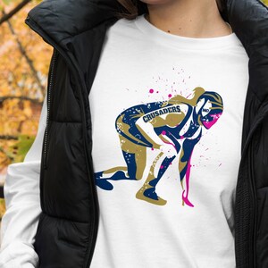 Può includere: Maglietta bianca a maniche lunghe con una grafica di una persona in uniforme blu e oro con la scritta "Crusaders". La grafica è circondata da schizzi di vernice rosa.