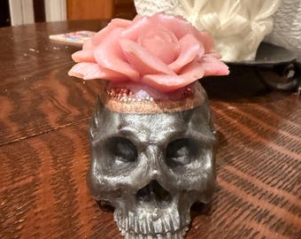 May include: A silver skull ornament with a pink rose on top. The rose is a detailed, multi-petaled design. The skull has intricate features and a metallic finish. This decorative item could be a candle or a unique home decor piece.