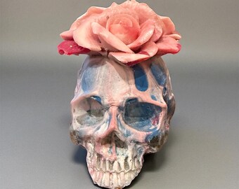 May include: A decorative skull sculpture featuring a marbled finish in shades of blue, pink, and white. A large, detailed pink rose is attached to the top of the skull, creating a contrast between the floral element and the bone structure.