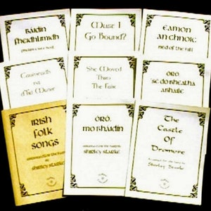 May include: A collection of nine white cards with black text, each featuring a different Irish folk song title. The cards are arranged in three rows of three. The cards are on a brown surface. The text on the cards includes:  'Ridin'  'Dust I Go Bound?' 'Eamon an chnoc' 'Coumcoth na d'll (Duter' 'She Doved There The Fak' 'ORO sc do nheatha arhalc' 'IRISH folk songs' 'ORO. mo shaidin' 'The Castle of Dromure'