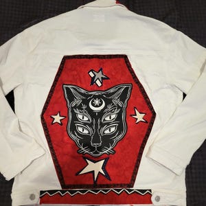 May include: White denim jacket with a large red and black cat graphic on the back. The cat has multiple eyes and is surrounded by stars. The jacket has a button-up front and long sleeves.