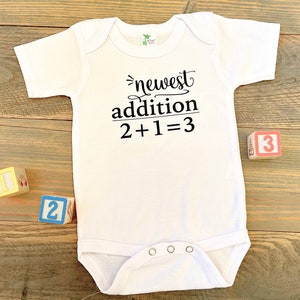 May include: White baby onesie with black text that reads "newest addition 2+1=3".  Three wooden blocks with the numbers 1, 2, and 3 are on a wooden surface.