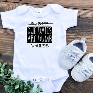 May include: White baby onesie with black text that reads "May 25, 2023 Due Dates Are Dumb April 11, 2023".